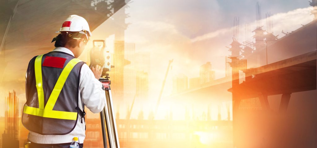 Imagem ilustrando as ações de um engenheiro civil, simulando um dos processos em vistoria cautelar de vizinhança. Na imagem: um homem branco e magro, usando um capacete de proteção com um colete cinza e amarelo neon por cima da camisa branca observa com um medidor na altura dos olhos; ao fundo, a montagem de prédios refletidos contra a luz do sol.